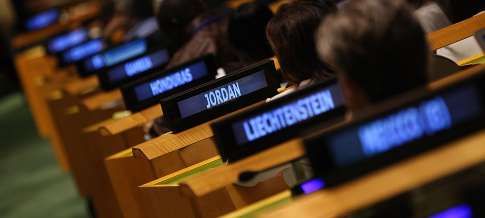 People sitting by desks with lit up monitors inside the UN Headquarters' Plenary room. Country names are written on the monitors.