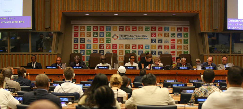 People sitting infront of the SDGs banner on the podium in the UN Plenary, infront of a room full of decision-makers