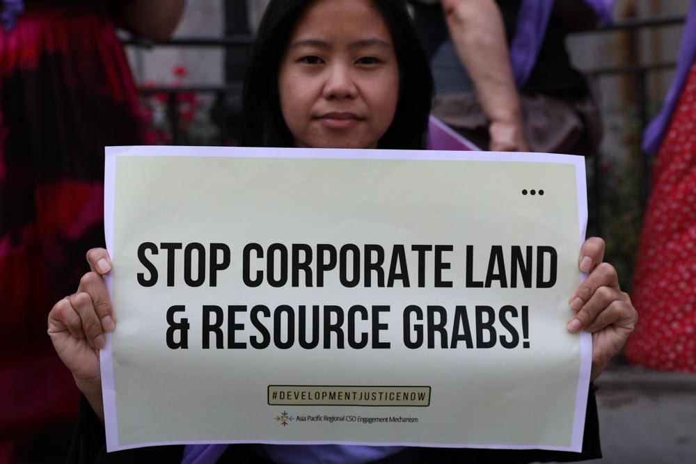A woman at a demonstration outside the UN looking straight into the camera, holding up a sign saying "Stop corporate land and resource grabs, development justice now"