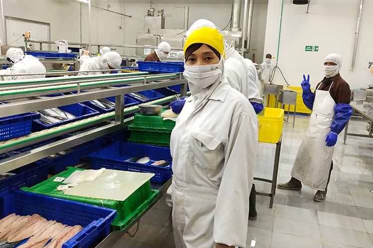 Workers at a fish factory wearing face masks. Woman at the forefront looking into the camera, man in the background showing the peace sign with his fingers.
