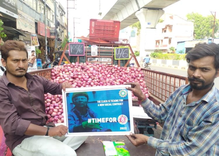 Two men sitting at the back of a truck holding up a sign in-between them saying "The time is ticking for a new social contract. Time for 8"