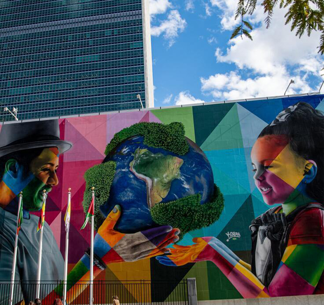 A photo of a UN wall that has art on it. A man holding a globe, passing it to a young girl. Lots of colours and geometric shapes